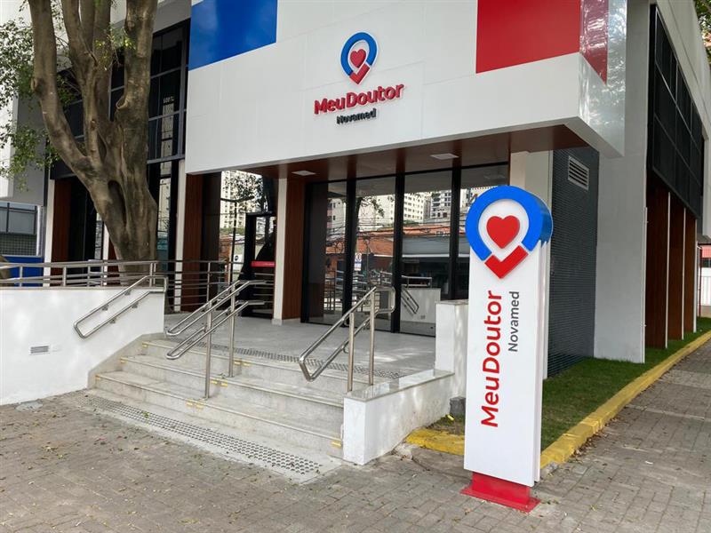  Fachada da unidade Meu Doutor  Novamed em tons de azul e vermelho  com o logotipo no topo. A entrada  possui portas de vidro, colunas  estruturais, escada de mármore claro  e rampa de acessibilidade. À direita, há  um totem branco da marca.